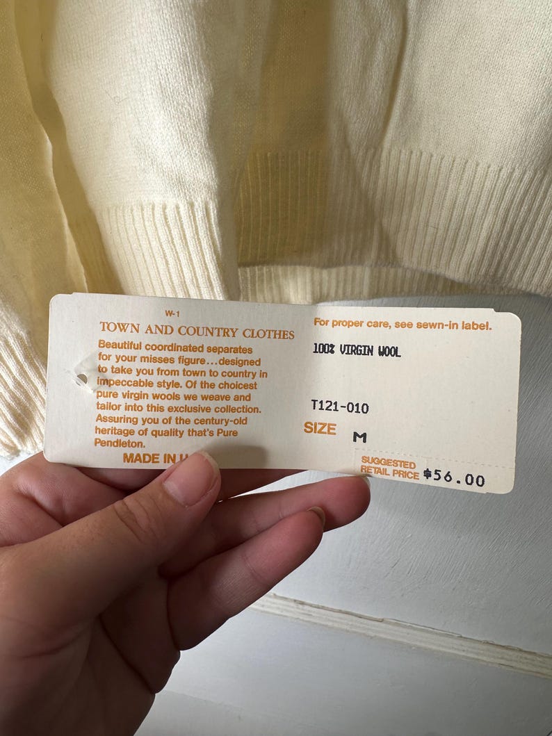 May include: Cream-coloured sweater with a ribbed hem and cuffs. A tag reads "TOWN AND COUNTRY CLOTHES" and "100% VIRGIN WOOL". The tag also indicates size M and a suggested retail price.