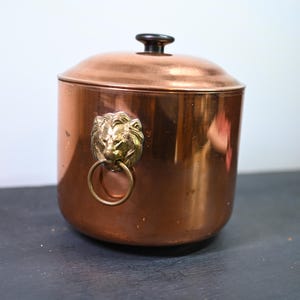 Pode incluir: Um balde de gelo de cobre com tampa e botão preto. O balde apresenta uma cabeça de leão de latão com uma alça de anel. O cobre tem um acabamento quente e reflexivo, e o balde tem formato cilíndrico.