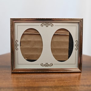 May include: A rectangular metallic picture frame with a copper-toned finish and a beaded edge. The frame features a cream-colored mat with two oval-shaped openings, each decorated with small, symmetrical brown scrollwork patterns at the top and bottom.