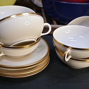 May include: A collection of vintage teacups and saucers. The cups and saucers are white with gold trim around the rims and handles. The teacups are stacked in pairs, and the saucers are stacked in sets of four.