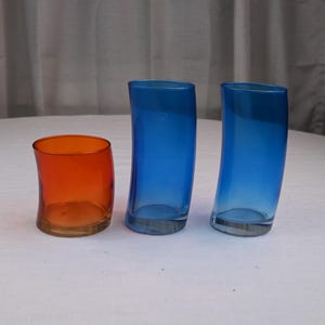 Può includere: Tre vasi di vetro colorati. Uno è arancione e gli altri due sono blu. I vasi hanno una forma curva e organica. Il vetro è trasparente, permettendo alla luce di passare. Questi vasi sono adatti per l'arredamento della casa.