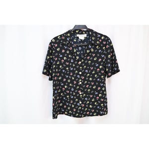 Puede incluir: Una camisa negra de manga corta con botones y un patrón repetitivo de pequeñas flores rosas, amarillas y azules intercaladas con formas rectangulares blancas. La camisa tiene un cuello clásico y una tapeta frontal con botones claros.