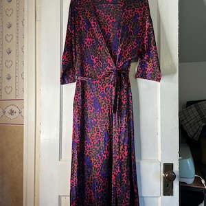 May include: A long wrap dress with a vibrant leopard print in shades of pink, purple, and brown hangs on a black hanger. The dress features a wrap design with a tie closure at the waist and three-quarter sleeves.