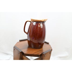 May include: A glossy, dark brown ceramic pitcher with a rounded body, a curved handle on the left side, and a light tan drip glaze accent along the top rim. The pitcher features a smooth, reflective surface and a distinct pouring spout.