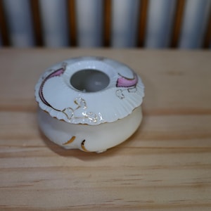 以下が含まれることがあります： 小さなオフホワイトのセラミック製の小物入れ。円形のデザインで、蓋にはピンクとゴールドのアクセント、スカラップの縁取りがあります。木製の表面に置かれています。