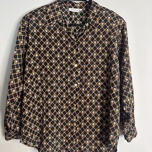 May include: Black button-down shirt with a repeating gold and white geometric pattern. The long-sleeved shirt has a collared neckline and gold-colored buttons. The shirt is hanging on a black hanger.