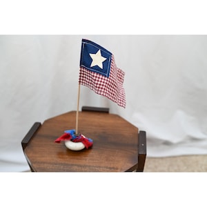 May include: A small fabric flag featuring a red and white gingham pattern with a blue square in the corner containing a white star. The flag is attached to a thin wooden pole, which is secured in a white circular base with red and blue fabric.