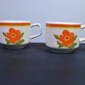 May include: Two ceramic mugs with a floral design. The mugs are off-white with an orange and yellow band around the rim. Each mug features a large orange flower with green leaves. The mugs have a handle.
