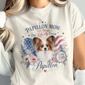 May include: Cream-colored t-shirt featuring a Papillon dog design. The graphic includes the text "Papillon Mom: Tiny Wings, Big Liberty Energy" with an American flag and floral accents. The design also includes the word "Papillon" and the number "250".