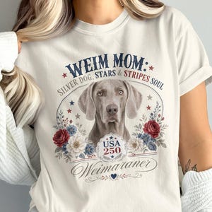 May include: Cream-colored t-shirt featuring a Weimaraner dog design. The graphic includes the text "WEIM MOM: SILVER DOG, STARS & STRIPES SOUL" with floral and star accents. The design also includes the text "USA 250" and "Weimaraner".