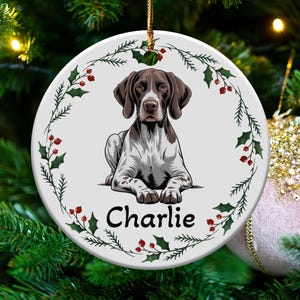 May include: A round, white Christmas ornament featuring a German Shorthaired Pointer dog illustration. The dog is brown and white, with the name "Charlie" below. The ornament is decorated with a holly and berry border, and is hanging on a Christmas tree.