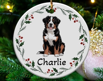 May include: A circular Christmas decoration with a Bernese Mountain Dog illustration. The dog is black, brown, and white, with the name "Charlie" beneath. The ornament is decorated with holly and red berries, suspended by a gold string.