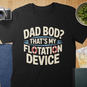 Peut inclure: T-shirt noir avec l'inscription "DAD BOD? THAT'S MY FLOTATION DEVICE" en blanc et bleu. Le motif comprend un graphique de bouée de sauvetage. Le t-shirt est présenté sur une surface en bois avec un appareil photo, un objectif et un bol de kiwis.