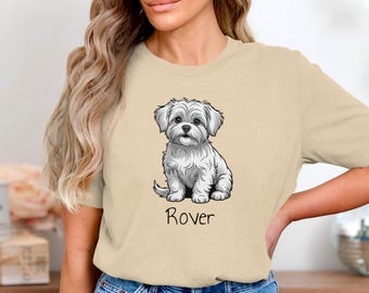 May include: A beige t-shirt featuring a detailed illustration of a small, fluffy white dog. The dog is sitting, with the word "Rover" printed in black script below. The shirt is a neutral color.