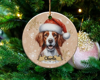 May include: A Christmas ornament featuring a brown and white dog wearing a red Santa hat. The ornament has a snowy background with gold snowflakes and the name "Charlie" written on a banner.