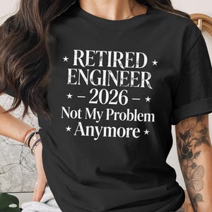 May include: Black t-shirt with the text "RETIRED ENGINEER - 2026 - Not My Problem Anymore" in white. The shirt has a crew neck and short sleeves. The text is centered on the front of the shirt.