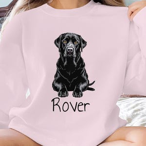 May include: Light pink sweatshirt featuring a black Labrador Retriever illustration with yellow eyes. The dog is sitting, and the name "Rover" is printed in black script below the dog. The sweatshirt is a crew neck style.