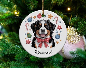 May include: A circular Christmas decoration with a Bernese Mountain Dog design, wearing a red bow tie, and festive embellishments. The dog's name, "Rascal," is written below. The ornament is suspended on a Christmas tree.