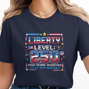 May include: Navy blue t-shirt with a pixelated graphic design. The design features the words "LIBERTY LEVEL: 250" in red, white, and blue, with stars, an American flag, and an eagle. The text "HIGH SCORE ACHIEVED" and "LEVEL 250 REACHED" are also included.