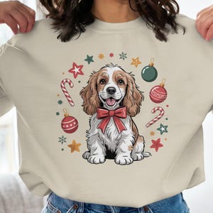 May include: Beige sweatshirt featuring a cartoon dog wearing a red bow tie, surrounded by Christmas ornaments, candy canes, and stars. The dog has brown and white fur and a happy expression. Festive holiday apparel.