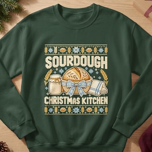 May include: A dark green crewneck sweatshirt featuring a graphic print with the text SOURDOUGH CHRISTMAS KITCHEN. The design includes a loaf of bread in a basket with a blue bow, a glass jar, and a folded cloth, surrounded by wheat stalks.