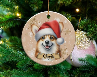 May include: A round Christmas ornament featuring a smiling corgi wearing a Santa hat. The ornament has a tan background with snowflakes and gold accents. A banner below the dog reads "Charlie".