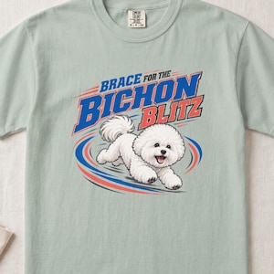 May include: A light sage green t-shirt featuring a graphic of a white Bichon Frise dog running. The text above the dog reads "BRACE FOR THE BICHON BLITZ" in bold blue and red lettering. The design includes blue and red motion lines surrounding the dog.