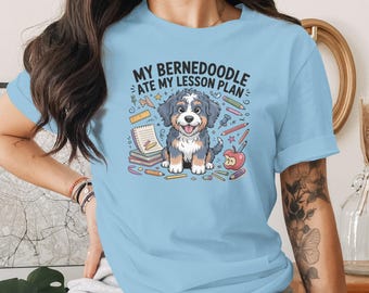 May include: Pale blue t-shirt with a cartoon Bernedoodle dog and the text "MY BERNEDOODLE ATE MY LESSON PLAN." The design includes pencils, an apple, and a notepad. The dog is sitting and smiling.