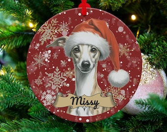Op de afbeelding: Ronde kerstversiering met een Whippet-hond met een kerstmuts. De versiering heeft een rode achtergrond met sneeuwvlokken en de naam "Missy" op een banner. Een feestelijke vakantieversiering.