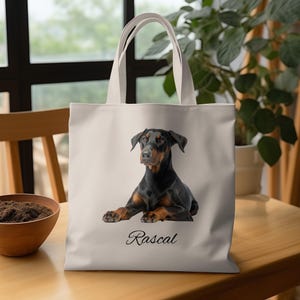 May include: A beige tote bag featuring a black and brown Doberman Pinscher dog lying down. The dog's name, "Rascal," is written in cursive below the image. The bag has two beige handles.