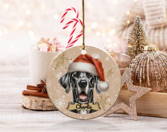 May include: A round ceramic Christmas ornament featuring a black Great Dane dog wearing a red Santa hat. The ornament has a white background with gold snowflakes and the name "Charlie" written on a banner.