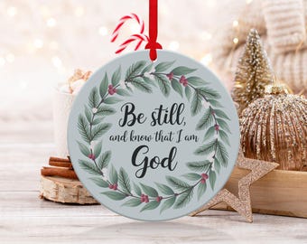 May include: A round Christmas bauble with a light blue background and a green wreath with red berries. The bauble has the text "Be still, and know that I am God" in black lettering.