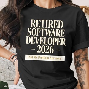 May include: Black t-shirt featuring the text "RETIRED SOFTWARE DEVELOPER - 2026 - Not My Problem Anymore" in an off-white, bold font. The shirt has short sleeves and a crew neck.