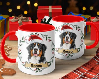 Peut inclure: Deux mugs en céramique blanche avec l'intérieur et les anses rouges. Chaque mug présente un bouvier bernois portant un bonnet de Père Noël, entouré d'une couronne. Le nom "Rosco" est affiché sur une bannière.