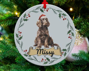 May include: A round Christmas ornament featuring a brown dog wearing a Santa hat. The dog is centered on a light gray background, surrounded by a wreath of holly and berries. The name "Missy" is displayed on a banner below the dog.