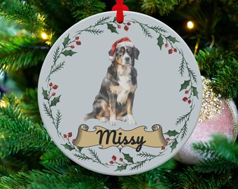 May include: Circular Christmas tree decoration with a dog wearing a Santa hat. The dog is black, brown, and white. The ornament has a light grey background with a holly and berry border. The name "Missy" is written on a banner.