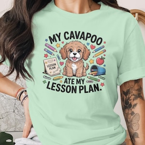 May include: A light green t-shirt featuring a cartoon Cavapoo dog with the text "MY CAVAPOO ATE MY LESSON PLAN." The design includes pencils, an apple, and a lesson plan. The shirt is made of a soft material.