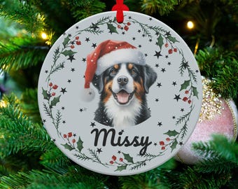 May include: A round Christmas ornament featuring a Bernese Mountain Dog wearing a Santa hat. The ornament has a light gray background with a holly and star border. The dog's name, "Missy," is written in cursive.