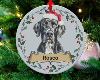 Peut inclure: Ornement de Noël rond avec un Grand Danois portant un bonnet de Père Noël. Le chien est noir et blanc sur un fond gris clair. L'ornement porte une bannière avec le nom "Rosco" et est entouré de houx et de baies.