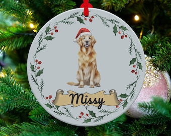 Puede incluir: Un adorno navideño de cerámica blanca con un borde de corona de acebo verde. Un perro golden retriever que lleva un gorro de Papá Noel rojo se sienta en el centro del adorno. El nombre del perro, "Missy", está escrito en una pancarta de rollo.