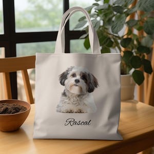 May include: A beige tote bag featuring a portrait of a fluffy dog with black and white fur. The dog's name, "Rascal," is written in cursive below the image. The bag has two white handles.