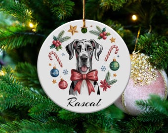 May include: A round Christmas ornament featuring a Great Dane dog with a red bow, surrounded by holiday decorations. The ornament has the name "Rascal" written on it. The ornament is hanging on a Christmas tree.