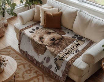 May include: A cream-colored throw blanket with a brown dog illustration and the name "Winston" on a scroll. The blanket has a fringe border and is draped over a white couch.