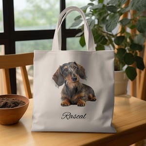 May include: A cream-coloured tote bag with a detailed illustration of a Dachshund dog. The dog has brown and black fur. The word "Rascal" is written in a cursive font below the dog. The bag has cream-coloured handles.