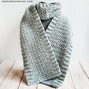 May include: A light gray crocheted scarf with a textured pattern. The scarf is folded and displayed on a wooden surface.