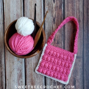 May include: A pink crocheted bag with a white border and a white strap. The bag is sitting on a wooden surface with two balls of yarn and a crochet hook.