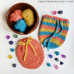 May include: Two crocheted drawstring bags, one in coral with yellow yarn ties, the other in turquoise, yellow, and coral stripes. Both bags are filled with dice.
