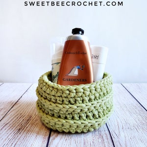 Może przedstawiać: Zielony koszyk na szydełku wypełniony różnymi artykułami toaletowymi, w tym butelką kremu do rąk Crabtree & Evelyn Gardeners, butelką mydła do kąpieli i białym tubką lotionu ochronnego 24H.