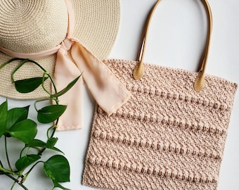 May include: A crocheted tote bag with brown leather handles. The bag is a light pink color and has a textured stitch pattern. The bag is sitting on a white surface next to a straw hat and a green plant.