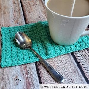 May include: A teal crochet coaster with a silver spoon resting on top. The coaster is sitting on a wooden surface with a white coffee mug containing coffee.